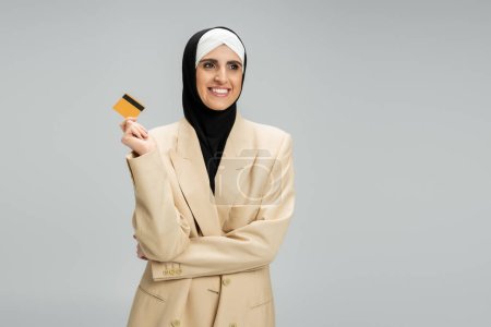 pleased middle eastern woman in muslim hijab and beige blazer holding credit card on grey