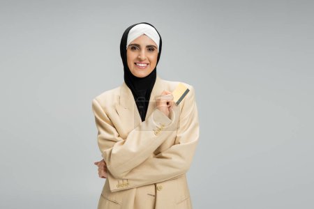 independent muslim businesswoman in beige blazer and hijab standing with credit card on grey
