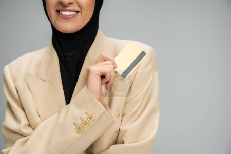 Téléchargez les photos : Vue recadrée d'une femme d'affaires musulmane souriante en blazer et hijab tenant une carte de crédit sur gris - en image libre de droit