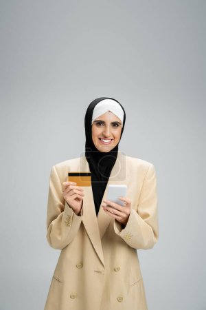 cheerful muslim businesswoman in hijab and trendy blazer holding smartphone and credit card on grey
