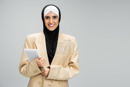 mujer de negocios musulmana optimista y de moda con tableta digital mirando a la cámara en gris