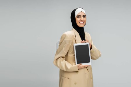 glückliche muslimische Geschäftsfrau in Hijab und Blazer zeigt digitales Tablet mit leerem Bildschirm auf grau
