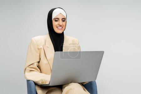 satisfied businesswoman in suit and hijab networking on laptop in armchair on grey