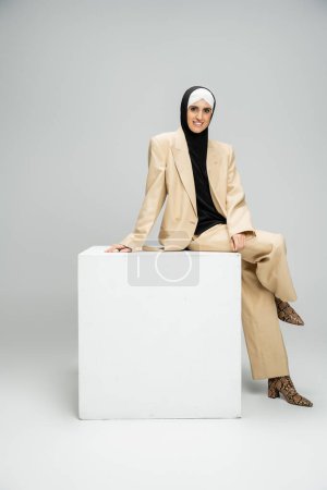 Foto de Alegre musulmana mujer de negocios en traje de moda y hijab sentado en cubo blanco en gris - Imagen libre de derechos