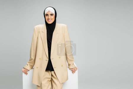 Foto de Feliz musulmana mujer de negocios en traje beige y hijab sonriendo a la cámara cerca de cubo blanco en gris - Imagen libre de derechos