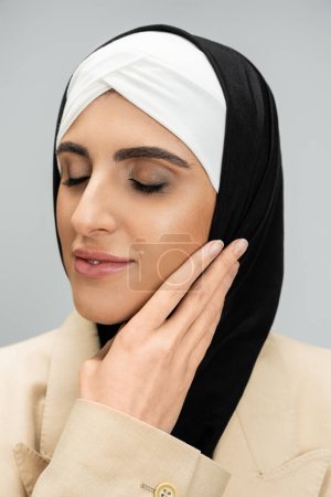 portrait of delighted and elegant muslim businesswoman with closed eyes touching face on grey