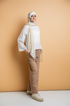 femme musulmane moderne en foulard, chemisier blanc, pantalon beige et bottes en cuir debout sur beige