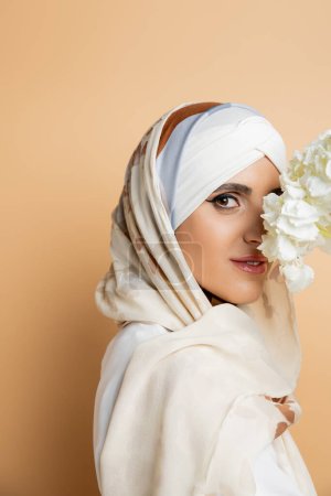 encantadora mujer musulmana en pañuelo para la cabeza oscureciendo la cara con flor blanca y mirando a la cámara en beige