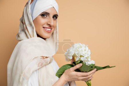 pleased muslim woman in blouse and headscarf, with hydrangea flower, looking at camera on beige