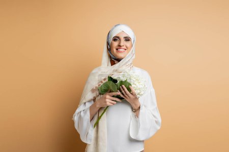 overjoyed muslim woman in blouse and headscarf holding hydrangea flower, looking at camera on beige