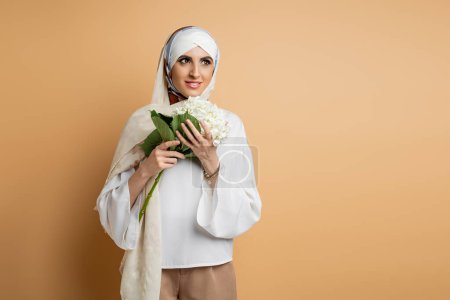 schöne muslimische Frau in seidenem Kopftuch und Bluse stehend mit weißen Hortensienblüten auf beige
