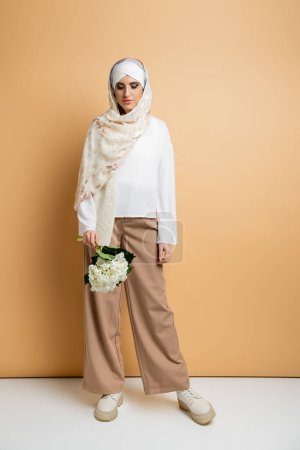 mujer musulmana moderna en pañuelo de seda y elegante atuendo casual posando con flor blanca en beige