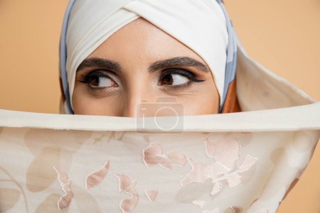 mujer musulmana elegante con maquillaje que oscurece la cara con un elegante pañuelo para la cabeza y mirando hacia otro lado en beige