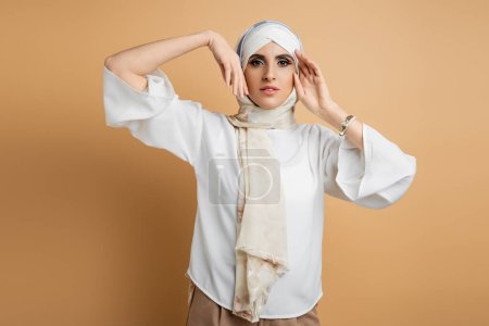 elegante Muslimin in weißer Bluse und seidenem Kopftuch posiert mit gesichtsnahen Händen auf beige