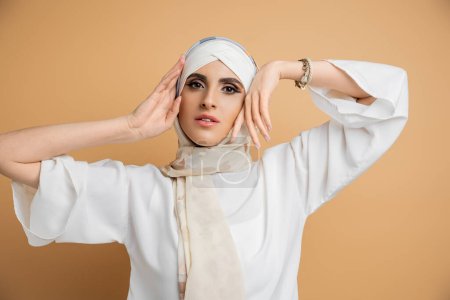 graceful muslim woman in elegant attire holding hands near face and looking at camera on beige