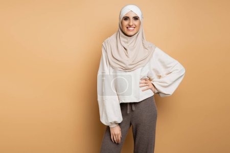 joyful muslim woman in hijab and trendy casual attire standing with hand on hip on beige