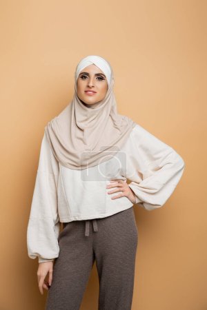 happy muslim woman in hijab and stylish casual clothes posing with hand on hip on beige