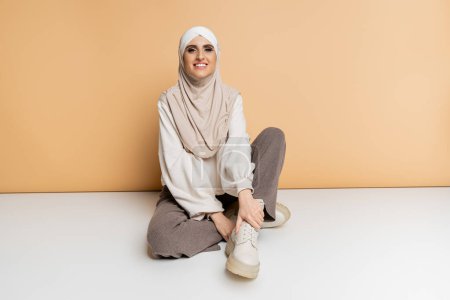 happy muslim woman in hijab and modern casual attire sitting on beige, full length, personal style