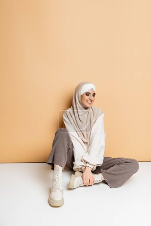 carefree muslim woman in fashionable casual outfit and hijab sitting on beige, full length