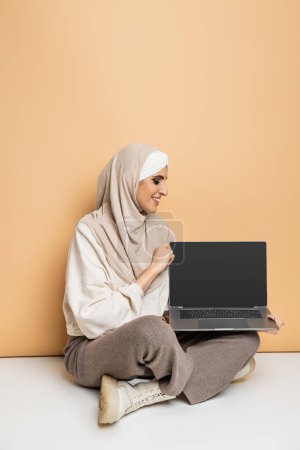 happy muslim woman in hijab and stylish casual attire sitting with laptop with blank screen on beige