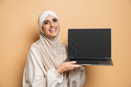 modern muslim woman in hijab holding laptop with blank screen and smiling at camera on beige