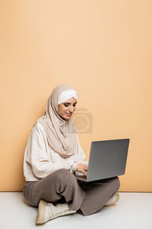 positive muslim woman in hijab and trendy casual outfit sitting and working on laptop on beige