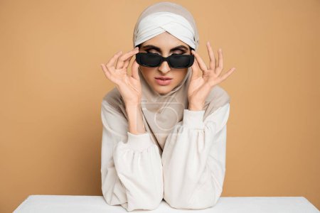 trendy muslim woman in hijab and sweatshirt sitting at white table, taking off sunglasses on beige