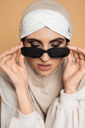 charmante femme musulmane en hijab enlevant des lunettes de soleil tendance sur beige, style personnel, individualité