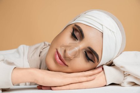 charming muslim woman with makeup wearing hijab and laying on white table with closed eyes on beige