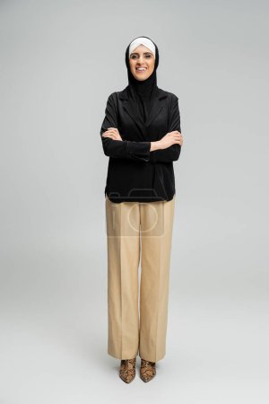 happy muslim businesswoman in hijab and stylish outfit posing with folded arms on grey, full length