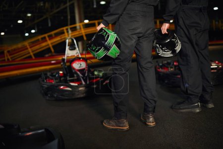 vista recortada de dos corredores de karts de pie cerca de los coches de carreras y la celebración de cascos, conductores de karts