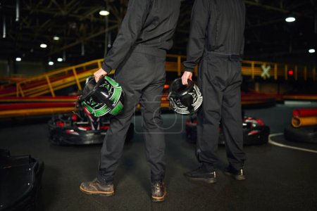 vista recortada de dos hombres de pie cerca de los coches de carreras y la celebración de cascos, el equipo de conductores de karts