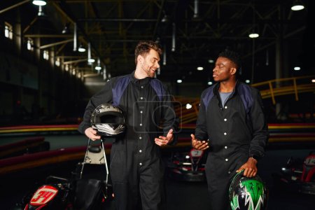 hombres multiculturales felices charlando dentro del circuito de kart, corredores de kart caminando y sosteniendo cascos