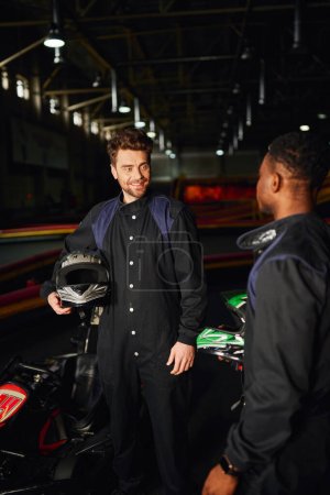 conducteur de kart joyeux tenant casque et regardant coureur afro-américain à l'intérieur du circuit de kart