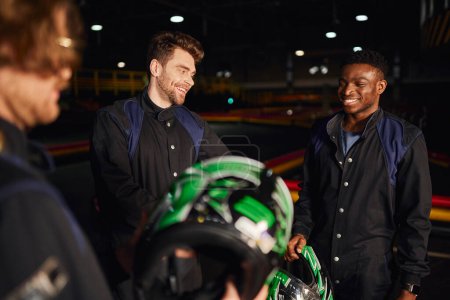 hombres multiculturales felices charlando dentro del circuito de kart, tres corredores de kart sosteniendo cascos
