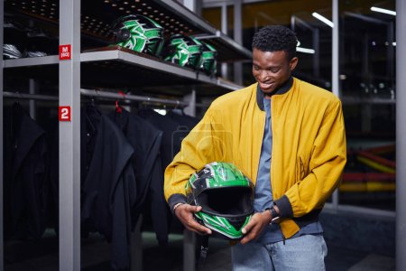 happy african american kart racer in yellow bomber jacket holding helmet, male driver, go-cart