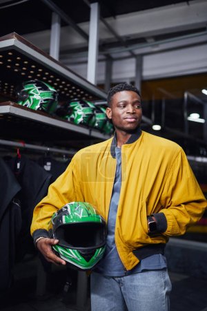 Photo for Confident african american kart racer in yellow bomber jacket holding helmet, male driver, go-cart - Royalty Free Image