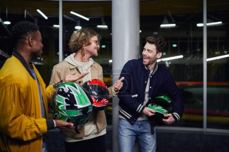 cheerful interracial male friends in jackets holding helmets, indoor racing track, karting concept