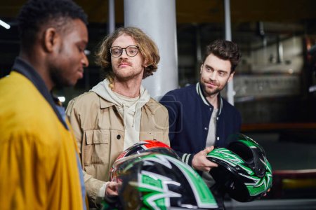 hombres con cascos mirando a amigo afroamericano dentro de pista de carreras cubierta, karting