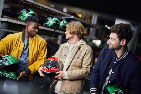 happy multicultural male friends in jackets holding helmets, indoor racing track, karting concept