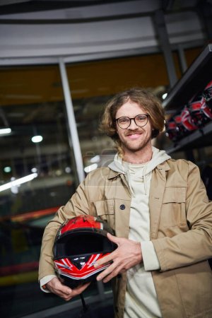 happy man in glasses holding helmet and looking at camera inside of indoor karting track, hobby