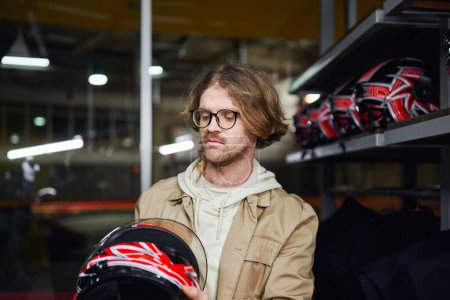 man in glasses looking at helmet inside of indoor karting track, motorsport and male hobby