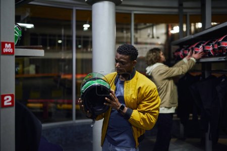 Foto de Africano americano hombre elegir casco para karting cerca amigo en fondo borroso, go-cart concepto - Imagen libre de derechos