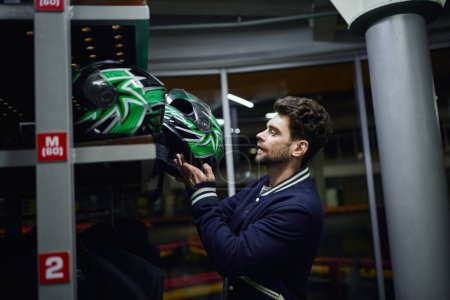 Foto de Hombre guapo elegir casco para karting dentro del vestuario de karting, concepto de automovilismo - Imagen libre de derechos