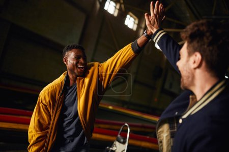 Téléchargez les photos : Excité homme afro-américain donnant haute cinq à un ami à l'intérieur de la piste de course de karting, la concurrence - en image libre de droit
