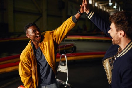 Téléchargez les photos : Excité homme afro-américain donnant haute cinq à un ami à l'intérieur de la piste de course de karting, gagner - en image libre de droit