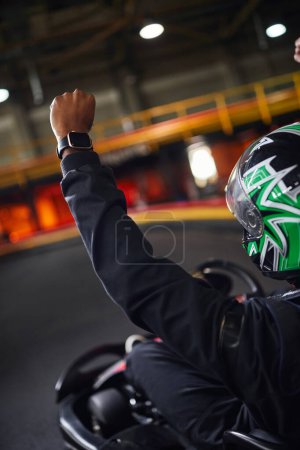 Go kart speed drive, excitado piloto afroamericano en casco levantando la mano y ganando carrera