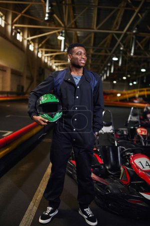 heureux homme afro-américain en vêtements de sport tenant casque et debout près de go-kart en piste de karting