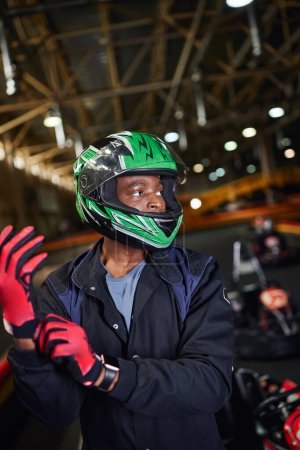 conductor de deportes de motor afroamericano en casco con guantes y de pie en pista de karting