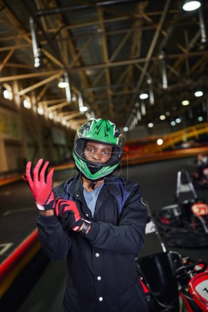ambitionierter afrikanisch-amerikanischer Motorsportfahrer mit Helm, Handschuhen und Stand in der Nähe von Go-Kart